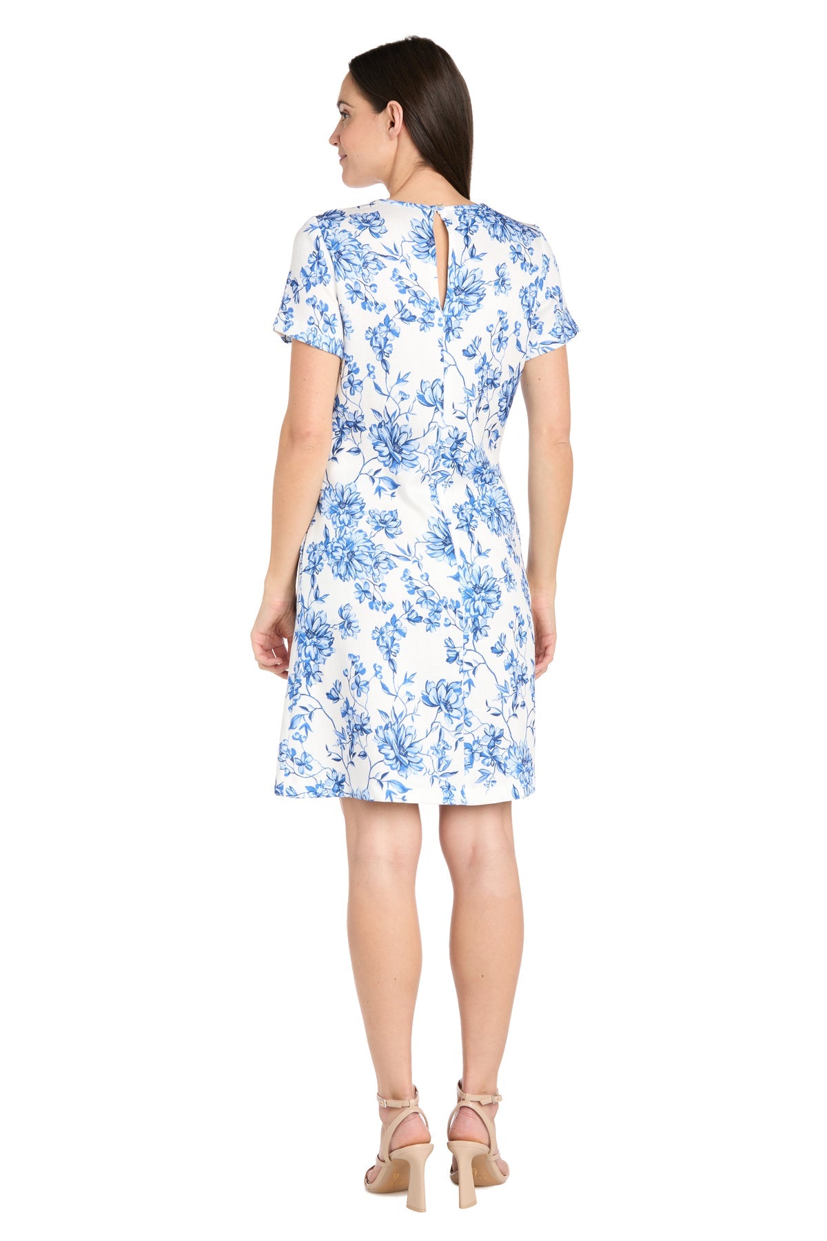 A woman stands facing away, wearing the R&M Richards 6674 short sleeve short party dress in blue and white floral print, paired with nude heeled sandals.